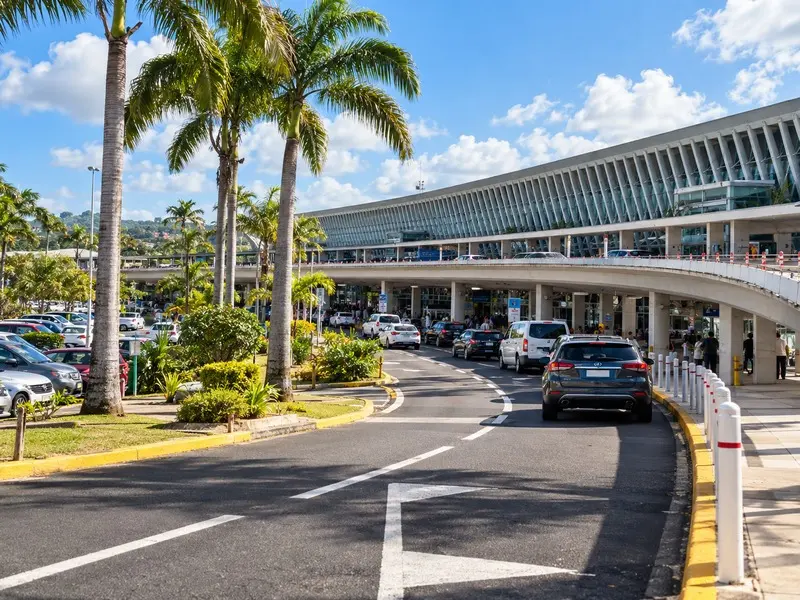 Entrée de l’aéroport Maryse-Condé en Guadeloupe avec voie de circulation, parking, palmiers et zone de dépose-minute des passagers aux Abymes.