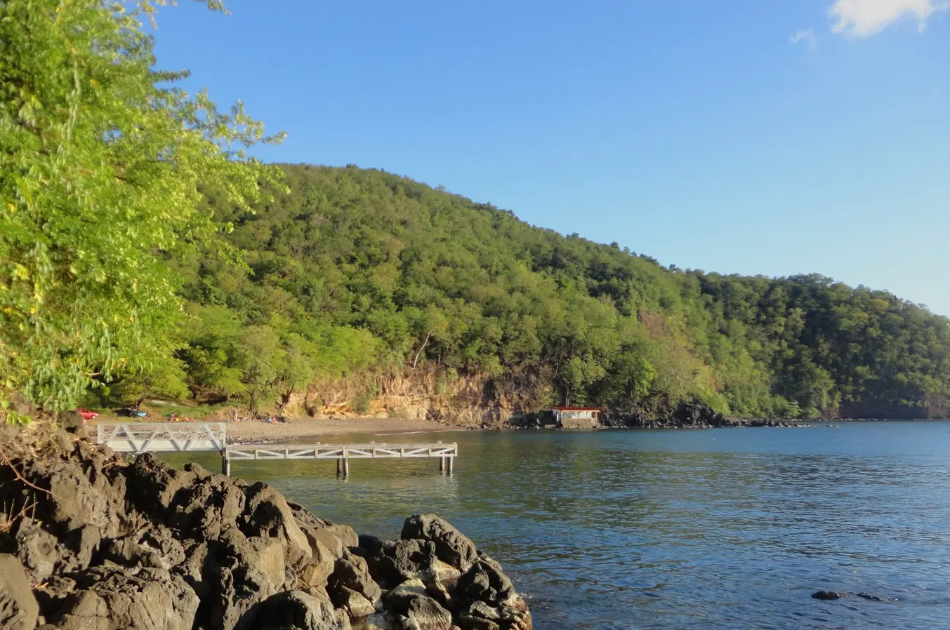 Vue panoramique depuis des rochers noirs de l'Anse à Sable à Bouillante en Guadeloupe, montrant un ponton en bois blanc s'avançant dans une mer calme, une petite plage nichée au pied d'une grande colline couverte d'une forêt dense sous un ciel bleu.