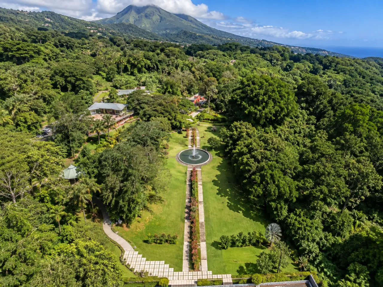 Domaine d’Émeraude en Martinique vu du ciel, avec ses jardins tropicaux, son bassin central, ses allées paysagères et la montagne Pelée en arrière-plan.