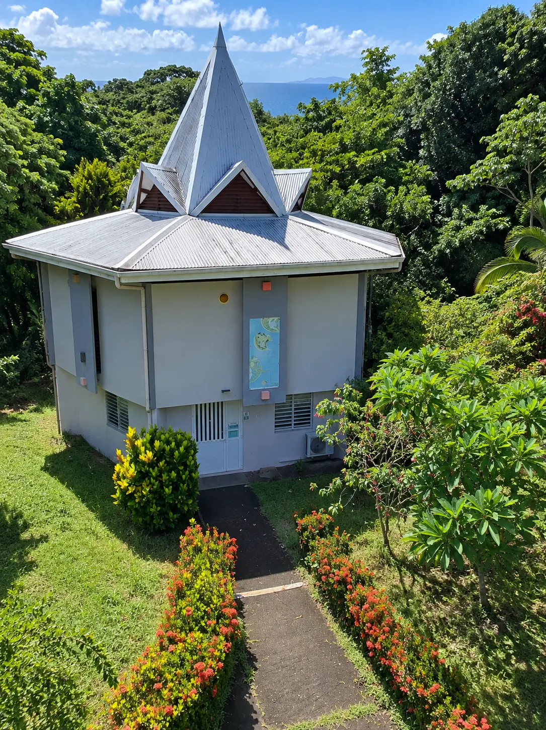 Édifice tropical en Martinique à l’architecture originale, avec toiture pyramidale métallique, jardin fleuri et végétation luxuriante en bord de mer.