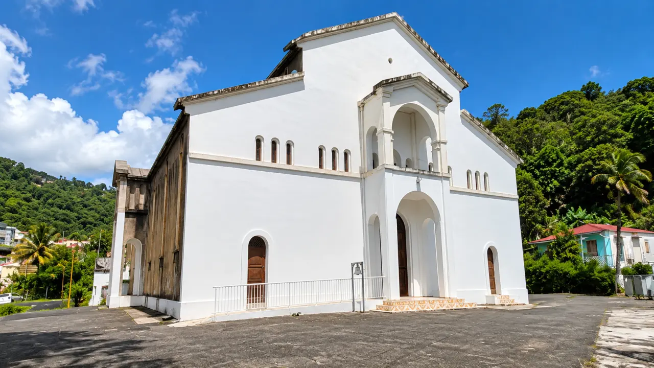 Église de Guadeloupe à flanc de montagne, architecture blanche à arcades et environnement tropical sous un ciel bleu.