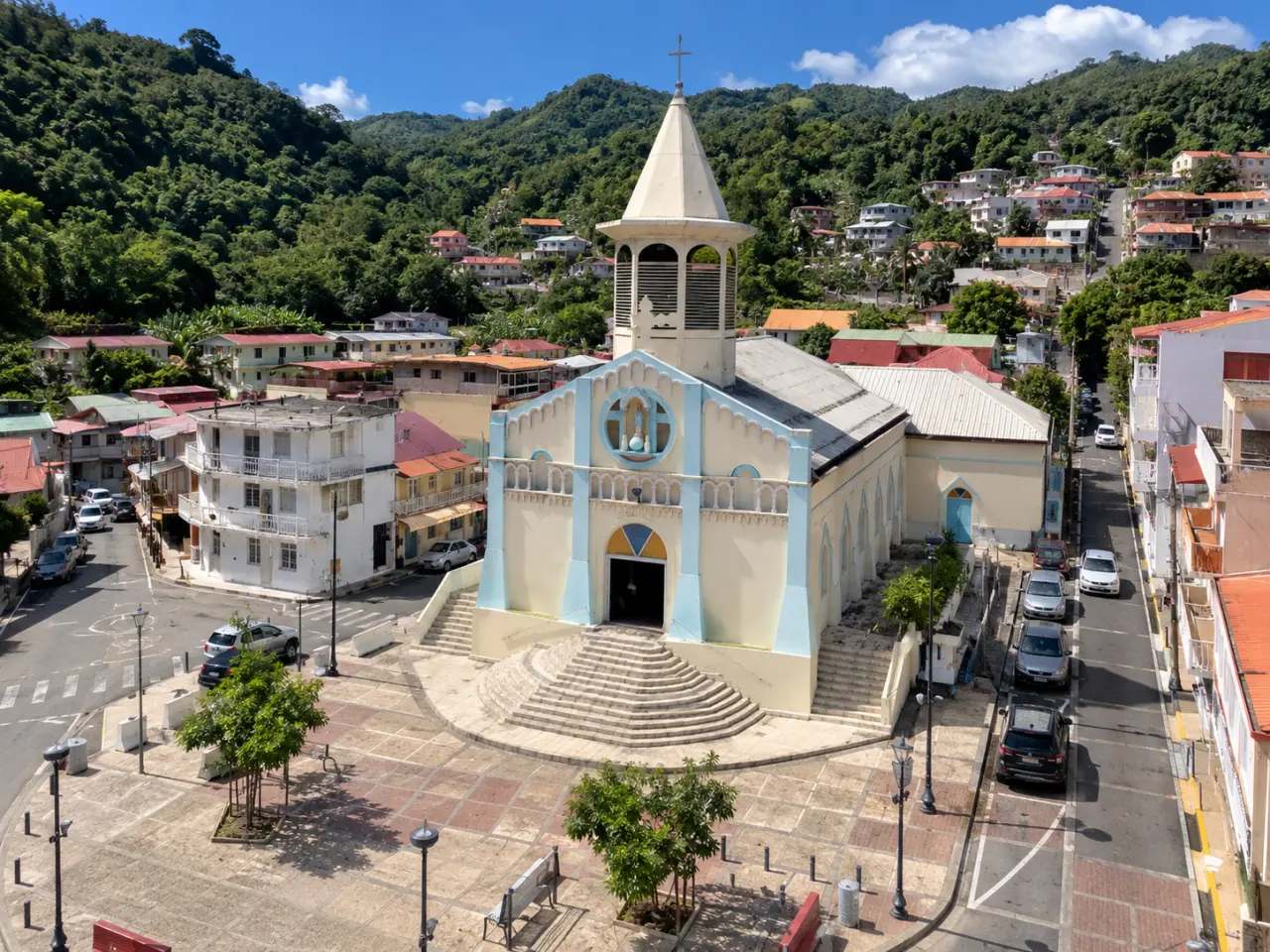 Église de Martinique vue du ciel, avec sa façade crème et bleu clair, son clocher à flèche et sa place entourée de maisons et de collines tropicales.