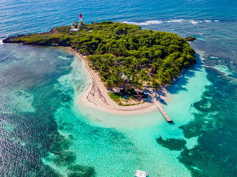 Photo aérienne de l'îlet du Gosier en Guadeloupe : lagon turquoise, plage de sable blanc en croissant et son phare rouge.