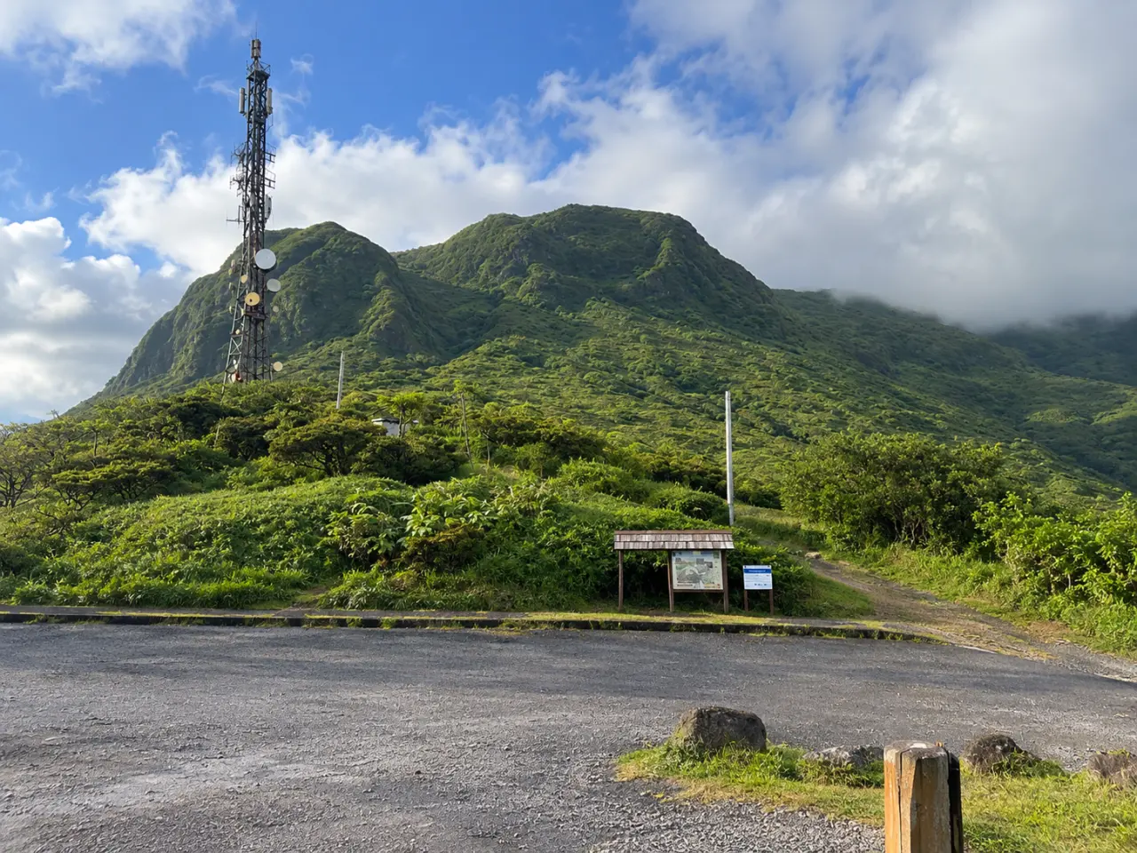Parking panoramique en Martinique au pied d’un relief montagneux verdoyant, avec une antenne de télécommunication et un sentier de randonnée.
