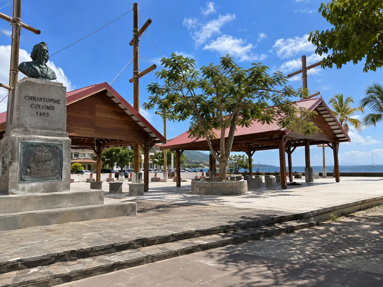 Place du débarquement de Christophe Colomb au Carbet en Martinique, avec ses pavillons en bois, sa statue commémorative et la mer des Caraïbes en arrière-plan.