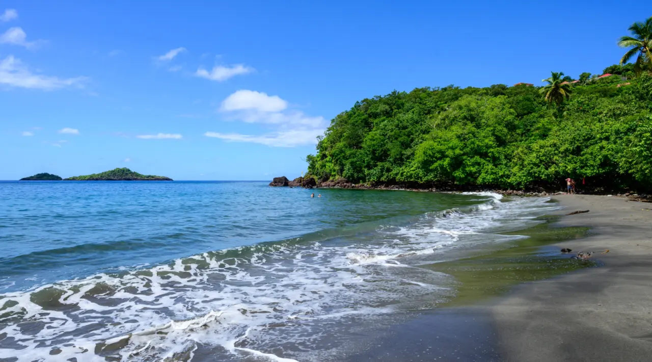 La plage de sable noir de Malendure à Bouillante en Guadeloupe, montrant de petites vagues à l'écume blanche touchant le bord, une colline verdoyante des arbres tropicaux à droite, les îlets Pigeon sur l'horizon de la mer des Caraïbes sous un ciel bleu.