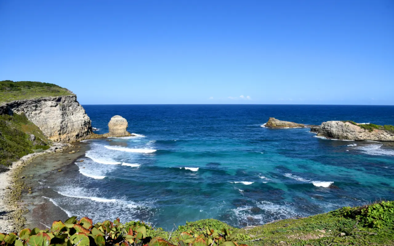 Vue panoramique de la Porte d'Enfer au Moule en Guadeloupe, montrant une baie aux eaux turquoise et agitées, entourée de falaises rocheuses sous un ciel bleu dégagé.