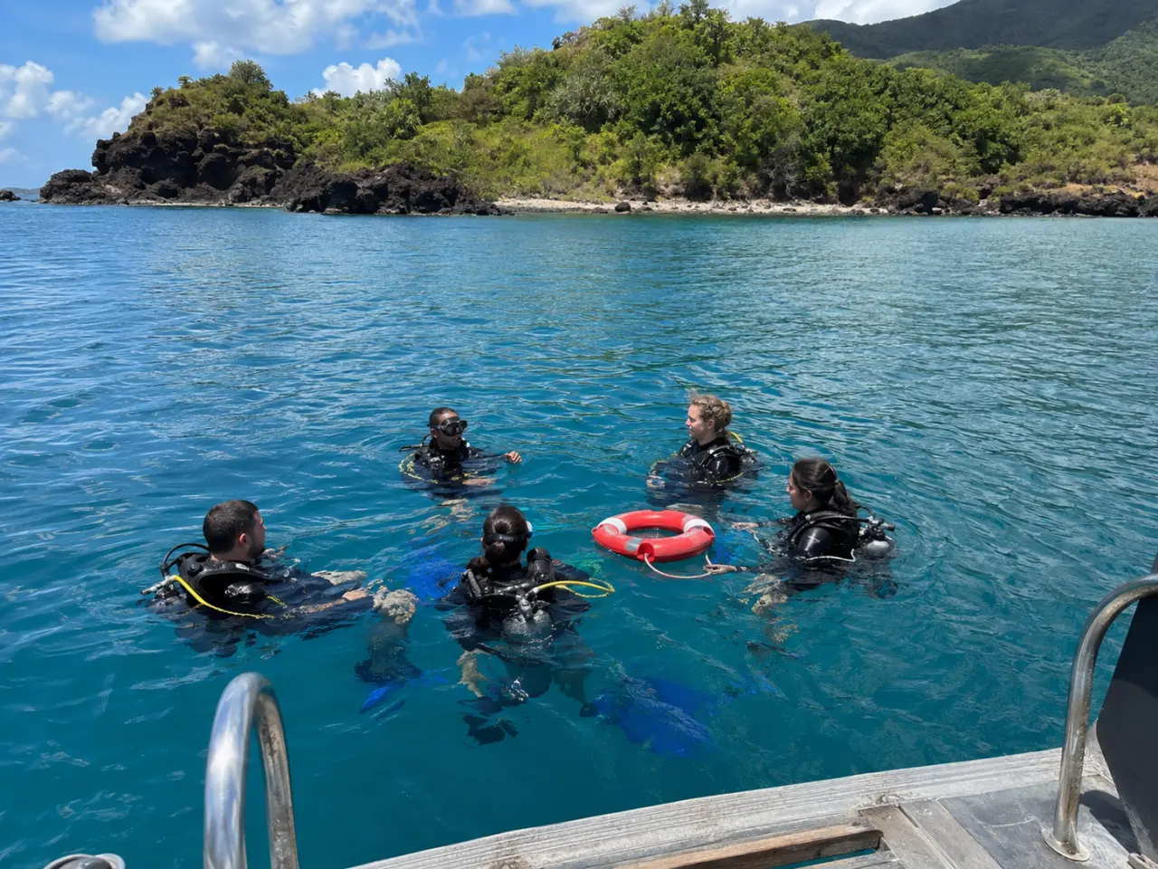 Groupe de plongeurs équipés de combinaisons et de bouteilles flottant en cercle dans l'eau turquoise de la Réserve Cousteau à Bouillante en Guadeloupe, à proximité d'une bouée de sauvetage rouge et blanche avec un îlet rocheux et boisé en arrière-plan.
