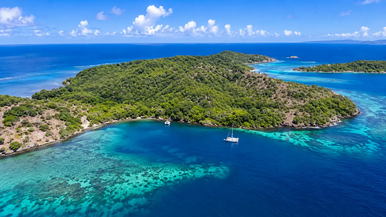 Vue aérienne d'une île tropicale de Guadeloupe entourée d'eaux turquoise, avec crique abritée, récifs coralliens et voiliers au mouillage.