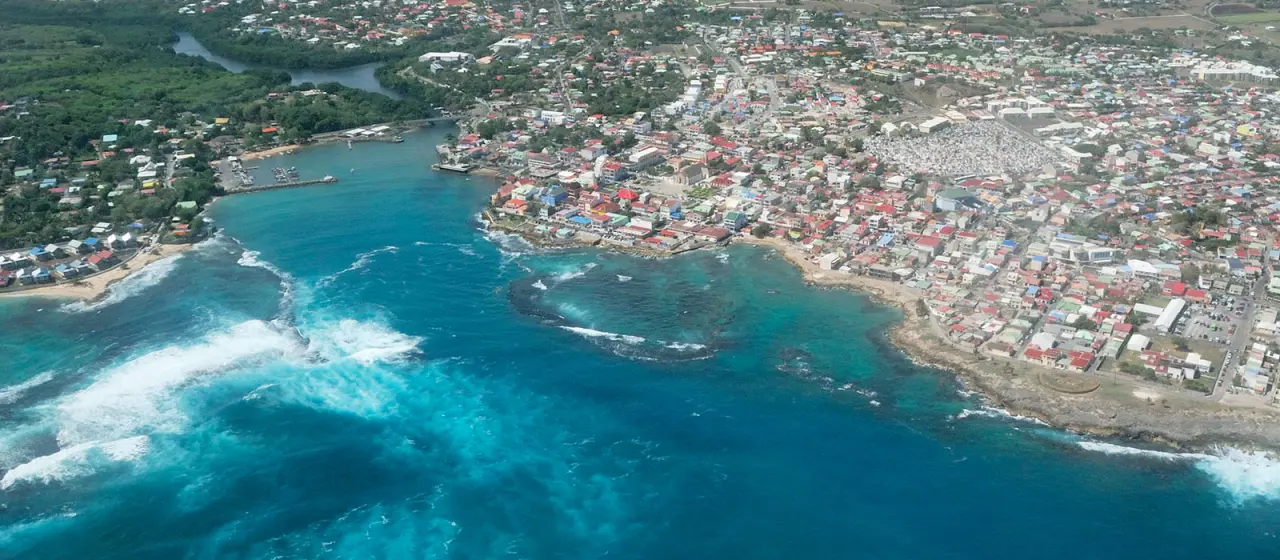 Vue aérienne du Moule en Guadeloupe : baie d'eau bleu turquoise, vagues à écume blanche et bourg littoral dense.