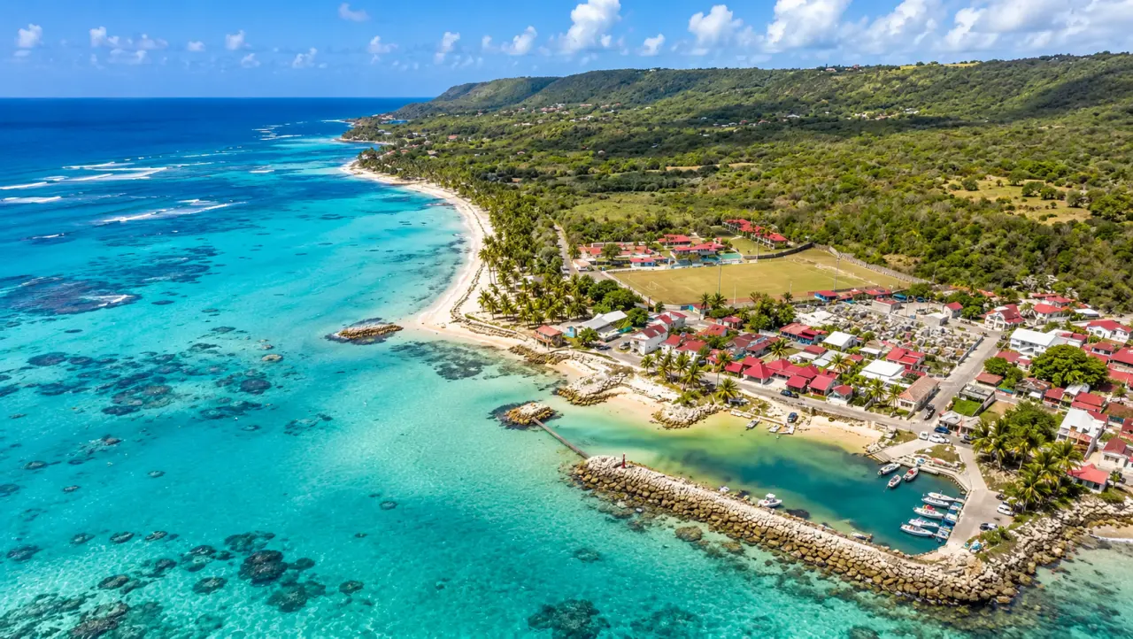 Vue aérienne de Capesterre-de-Marie-Galante en Guadeloupe avec plage, eaux turquoise, village côtier, port et collines verdoyantes.
