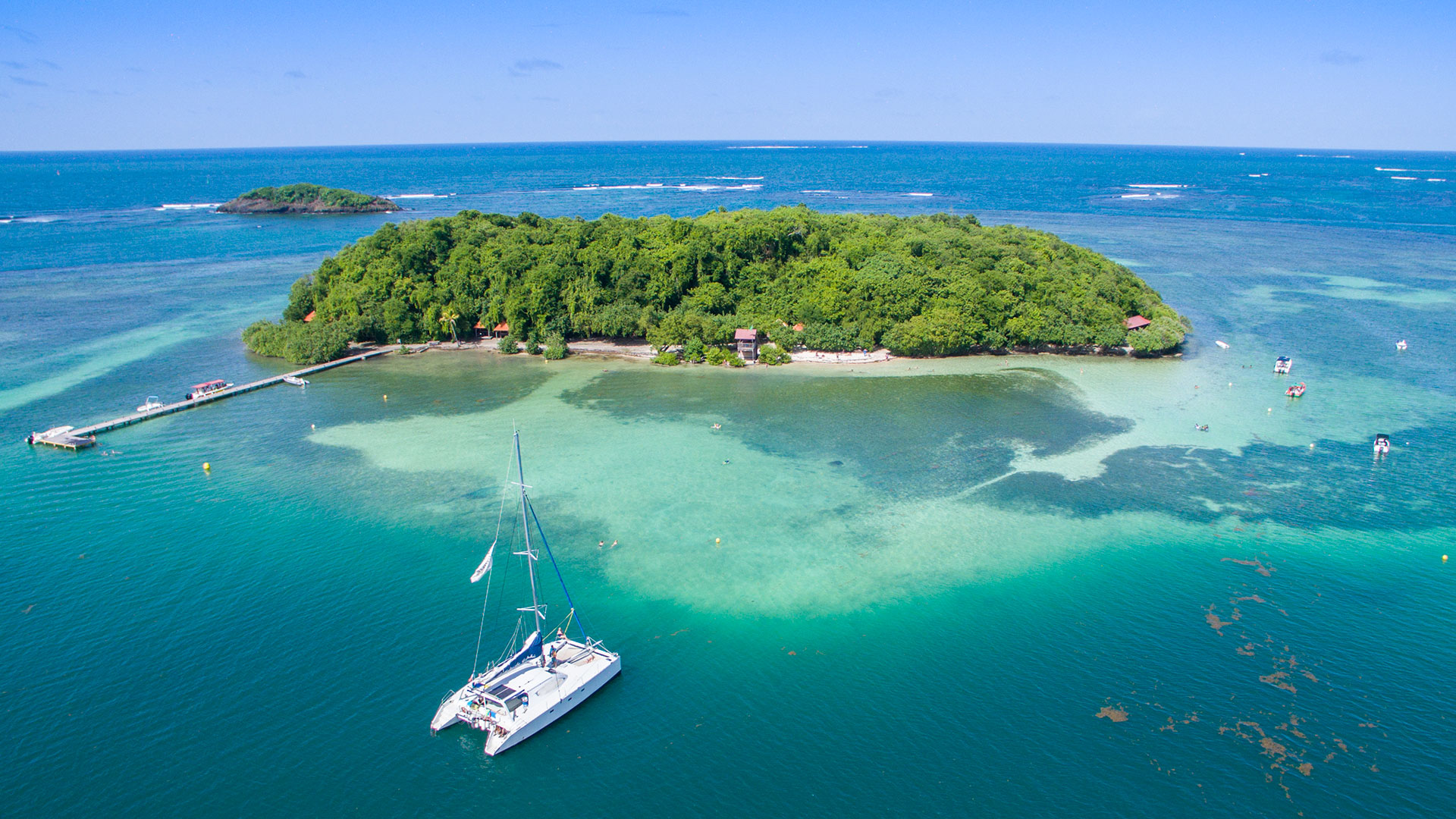 Vue aérienne de l'îlet Madame en Martinique : lagon turquoise transparent, catamaran blanc au mouillage et île boisée.