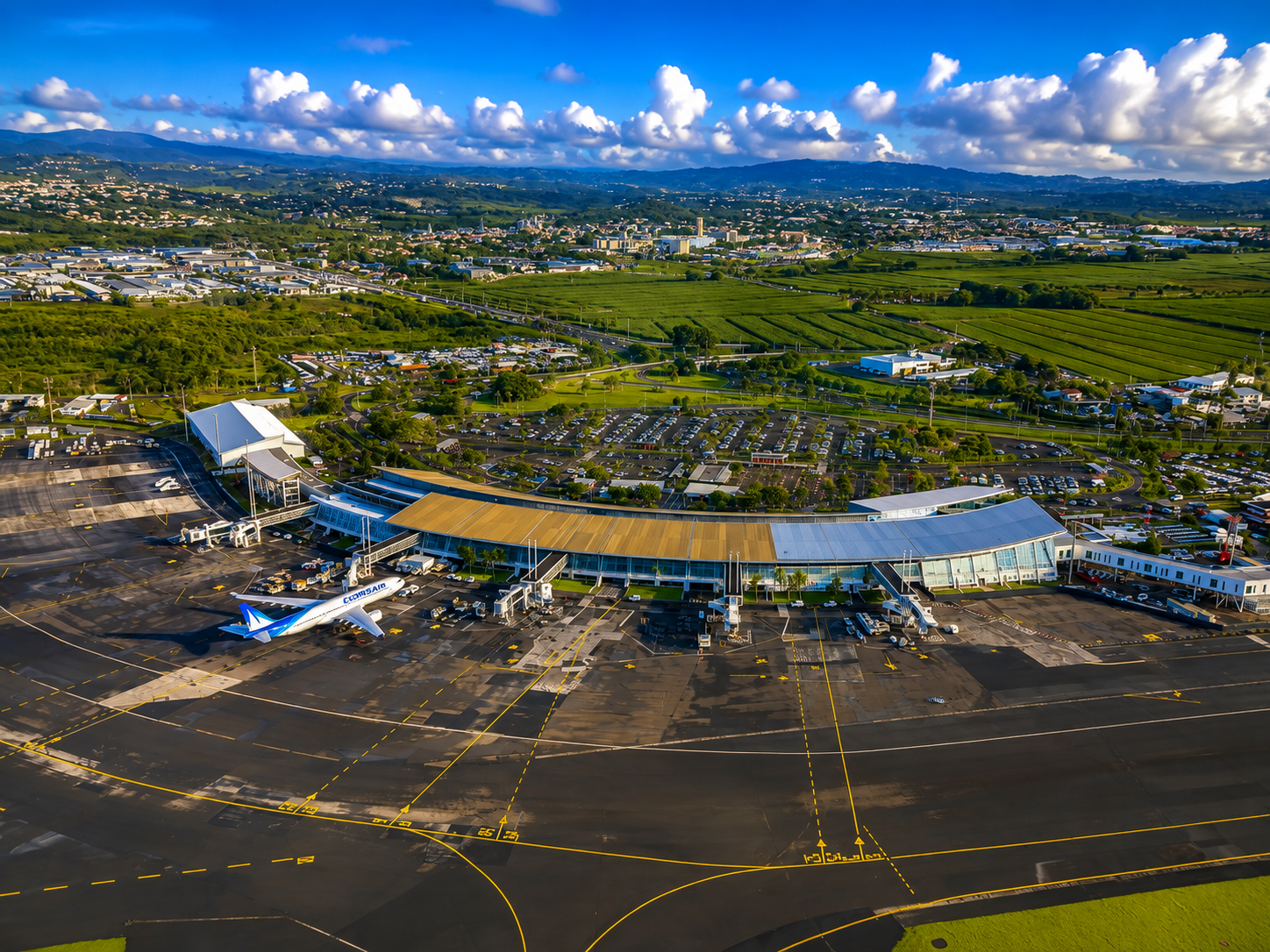 Vue aérienne de l'aéroport Martinique Aimé Césaire : terminal courbé ensoleillé, avion au sol et terres vertes au fond.