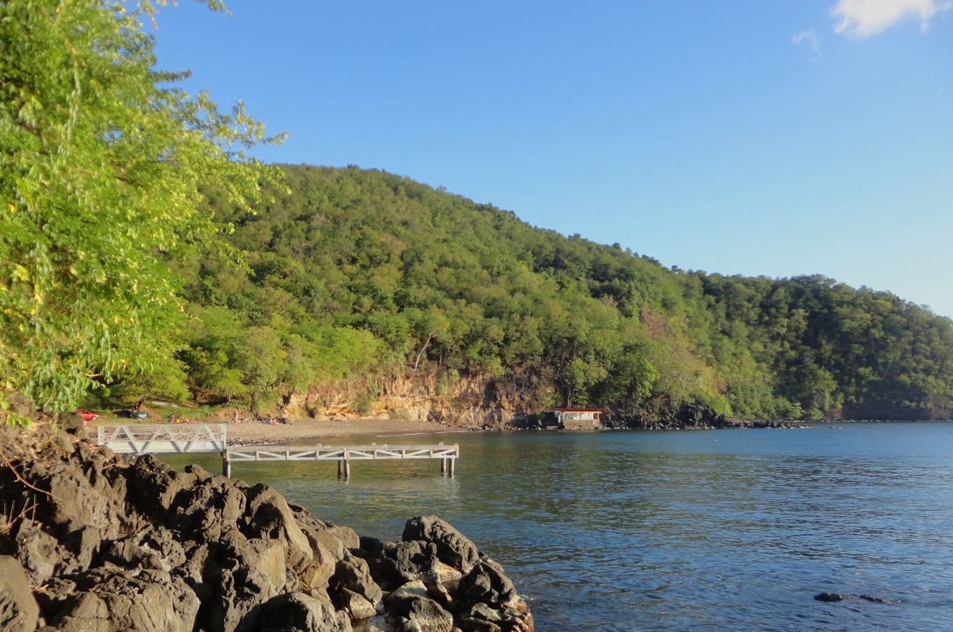 Vue panoramique depuis des rochers noirs de l'Anse à Sable à Bouillante en Guadeloupe, montrant un ponton en bois blanc s'avançant dans une mer calme, une petite plage nichée au pied d'une grande colline couverte d'une forêt dense sous un ciel bleu.