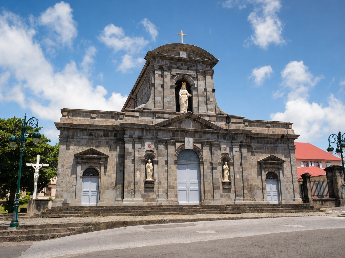 Façade en pierre de la Cathédrale Notre-Dame-de-Guadeloupe à Basse-Terre, affichant une architecture classique avec des statues de saints dans des niches et une statue de la Vierge Marie au sommet sous un ciel bleu.
