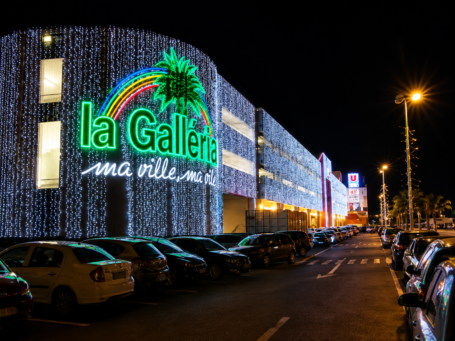 Centre commercial La Galleria en Martinique de nuit : façade illuminée de guirlandes, enseigne verte et parking voitures.
