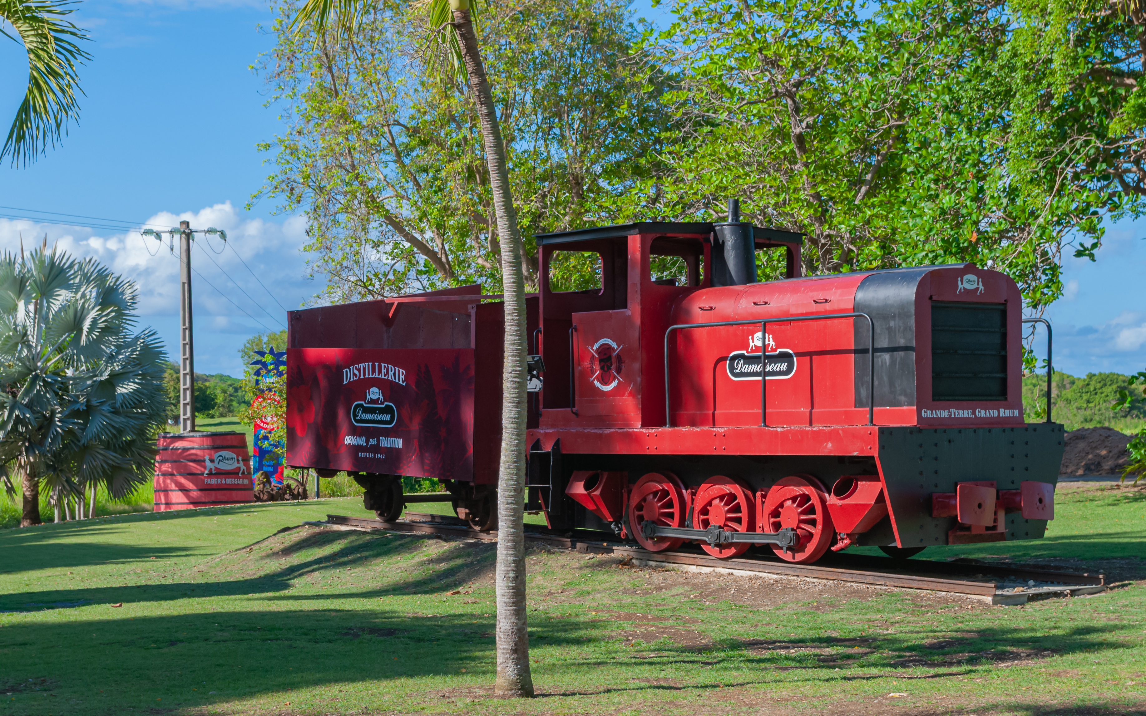 Ancien train sucrier rouge et noir exposé sur un rail à la distillerie Damoiseau au Moule, entouré de pelouse et d'arbres.