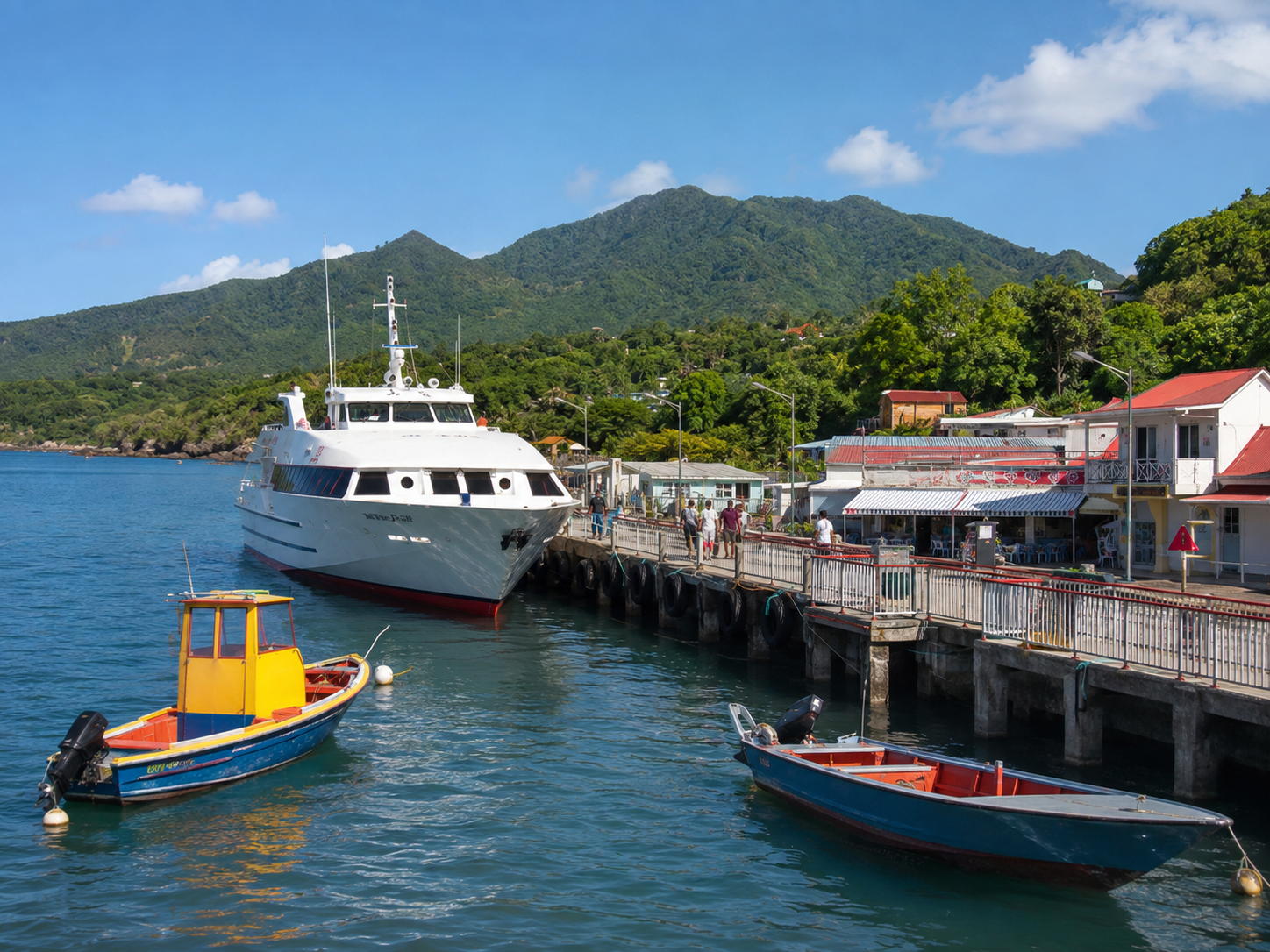 Vue de l'embarcadère de Trois-Rivières en Guadeloupe, montrant un grand ferry blanc accosté le long du quai, deux petites barques de pêche colorées flottant au premier plan, et des bâtiments côtiers aux toits rouges au pied de montagnes verdoyantes.