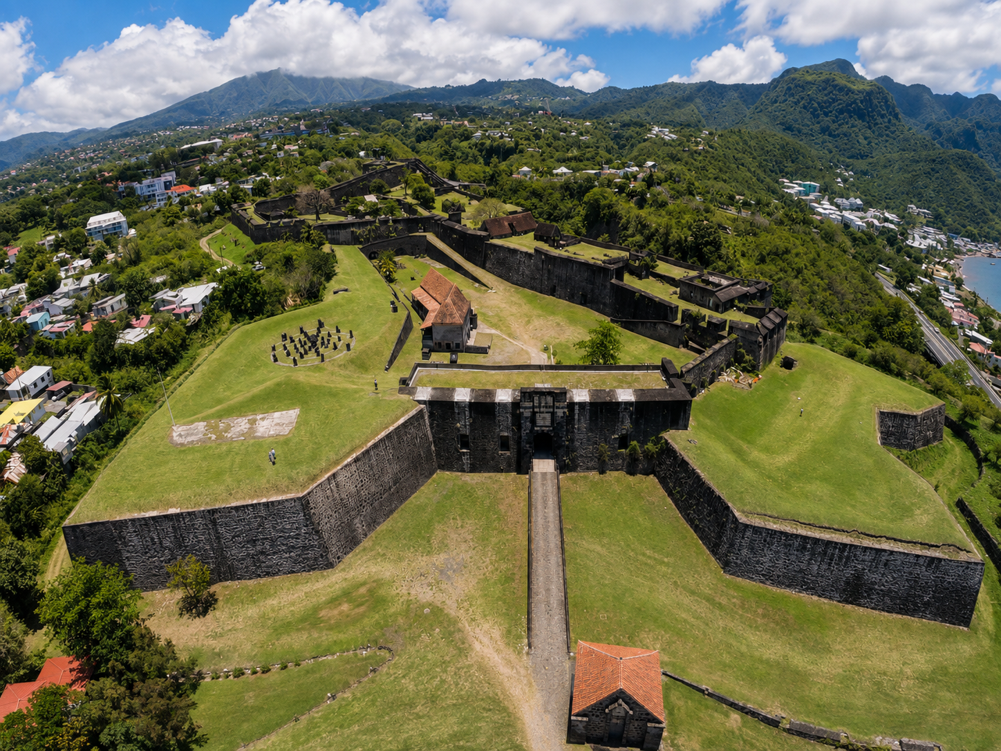 Vue aérienne du Fort Delgrès à Basse-Terre en Guadeloupe, mettant en valeur ses imposantes fortifications en pierre en forme d'étoile, ses bastions recouverts de pelouse verdoyante et les montagnes en arrière-plan sous un ciel nuageux.