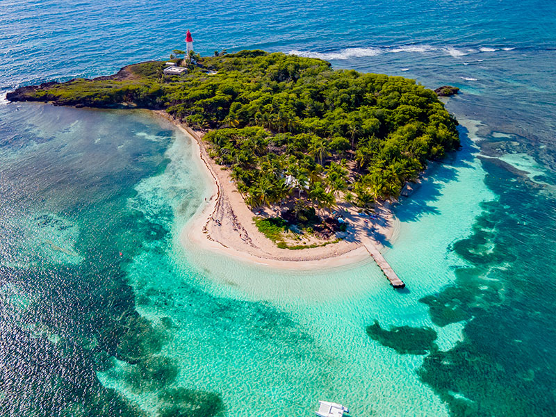 Photo aérienne de l'îlet du Gosier en Guadeloupe : lagon turquoise, plage de sable blanc en croissant et son phare rouge.