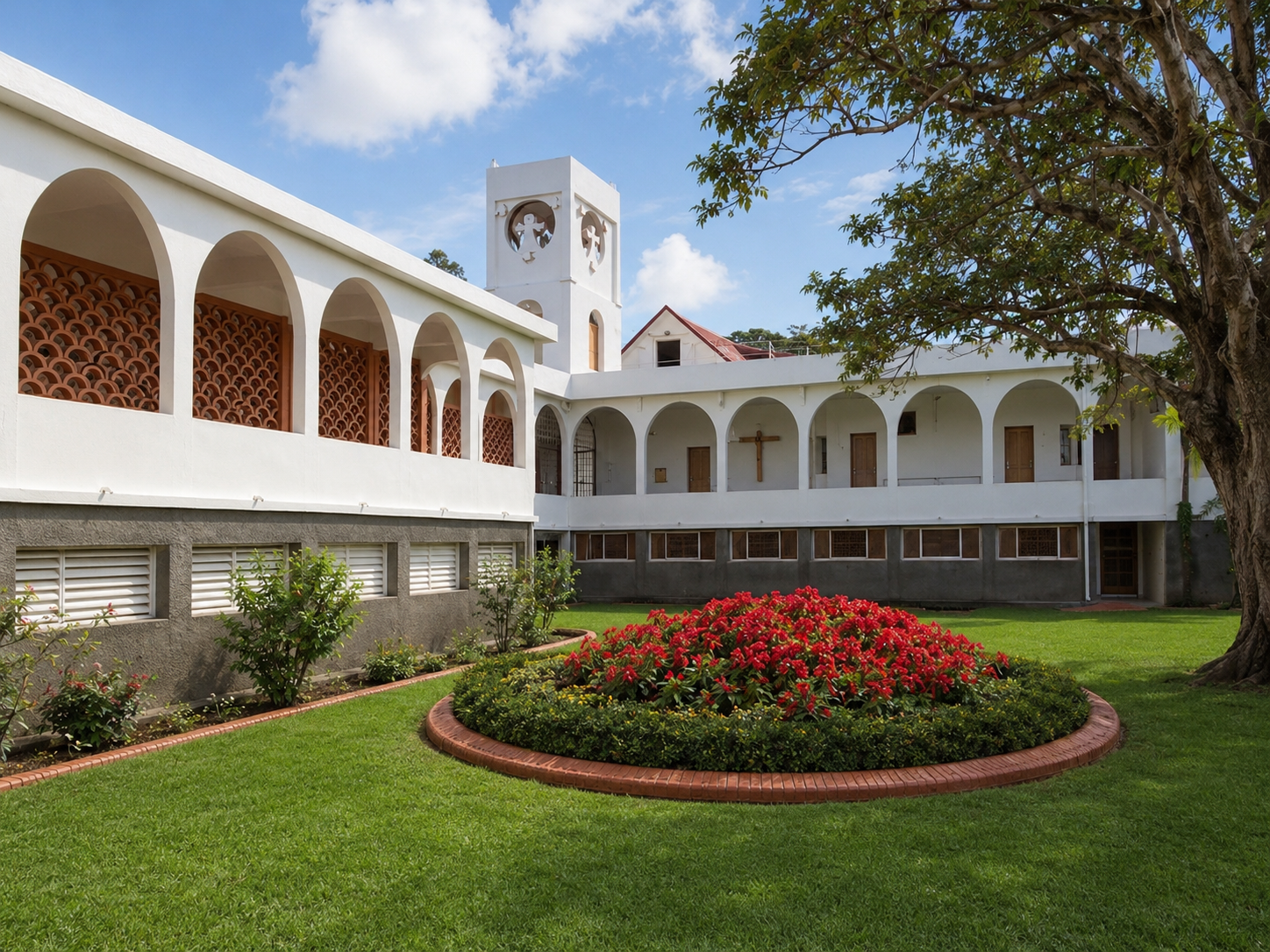 Cour intérieure du Monastère des Carmélites à Basse-Terre en Guadeloupe, montrant un jardin de pelouse bien entretenu avec un parterre circulaire de fleurs rouges au centre, entouré de bâtiments blancs à arcades et d'un clocher en arrière-plan.