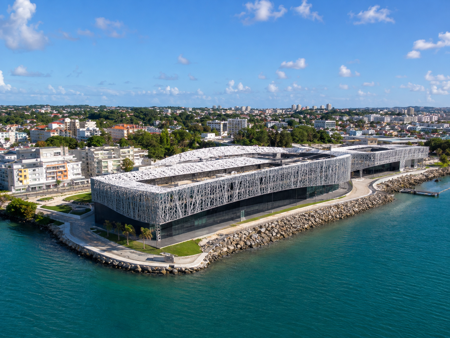 Vue aérienne du Mémorial ACTe à Pointe-à-Pitre en Guadeloupe, montrant son architecture moderne unique recouverte d'une structure en résille d'argent ajourée, implanté sur le front de mer en bordure d'une eau turquoise avec la ville en arrière-plan.