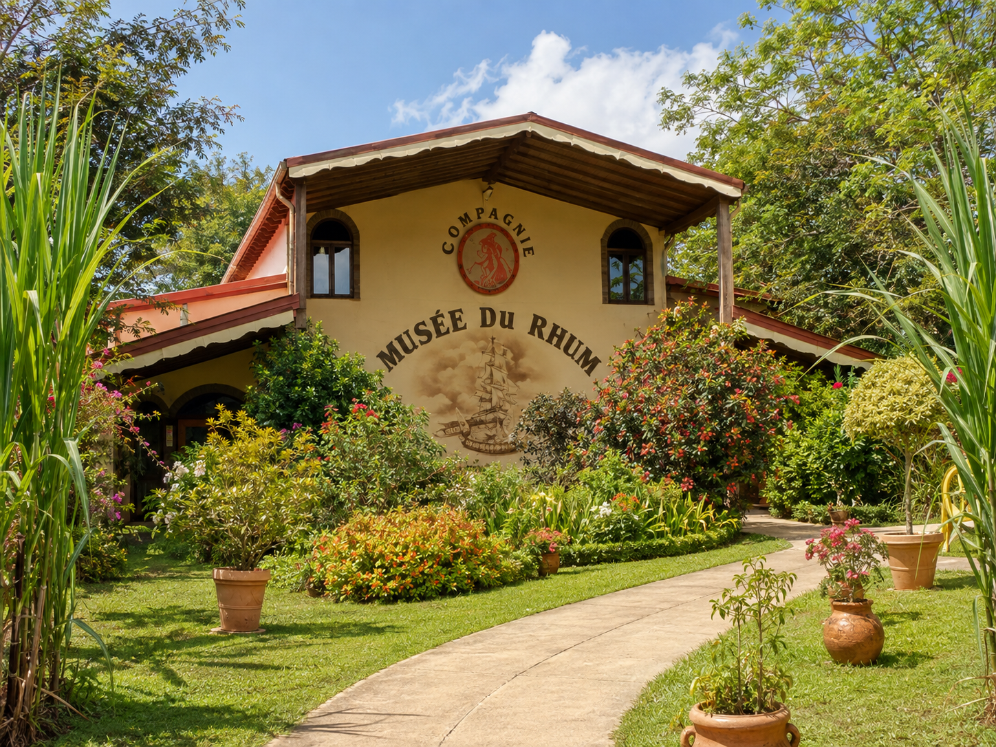 Façade extérieure du Musée du Rhum de la distillerie Reimonenq à Sainte-Rose en Guadeloupe, montrant un bâtiment de style créole jaune et marron, entourée d'un jardin tropical fleuri et de hautes tiges de canne à sucre au premier plan.