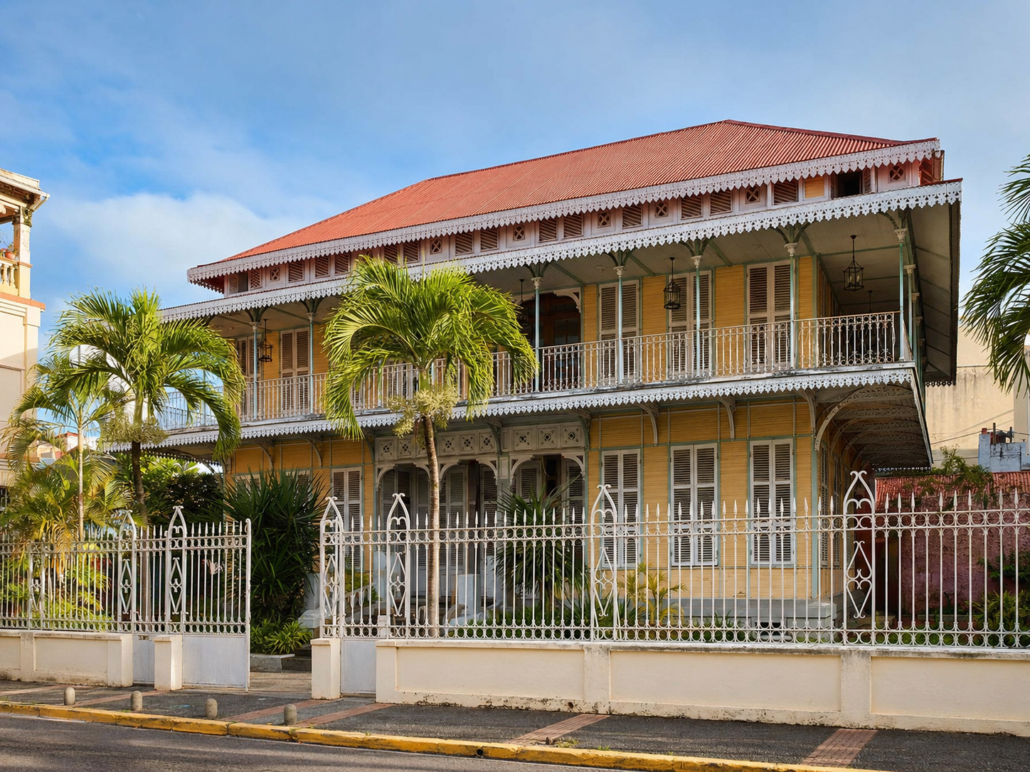 Façade d'une maison coloniale traditionnelle jaune à deux étages abritant le Musée Saint-John Perse à Pointe-à-Pitre en Guadeloupe, ornée de balcons en fer forgé, de frises en bois ciselé, et entourée d'une grille blanche avec des palmiers.