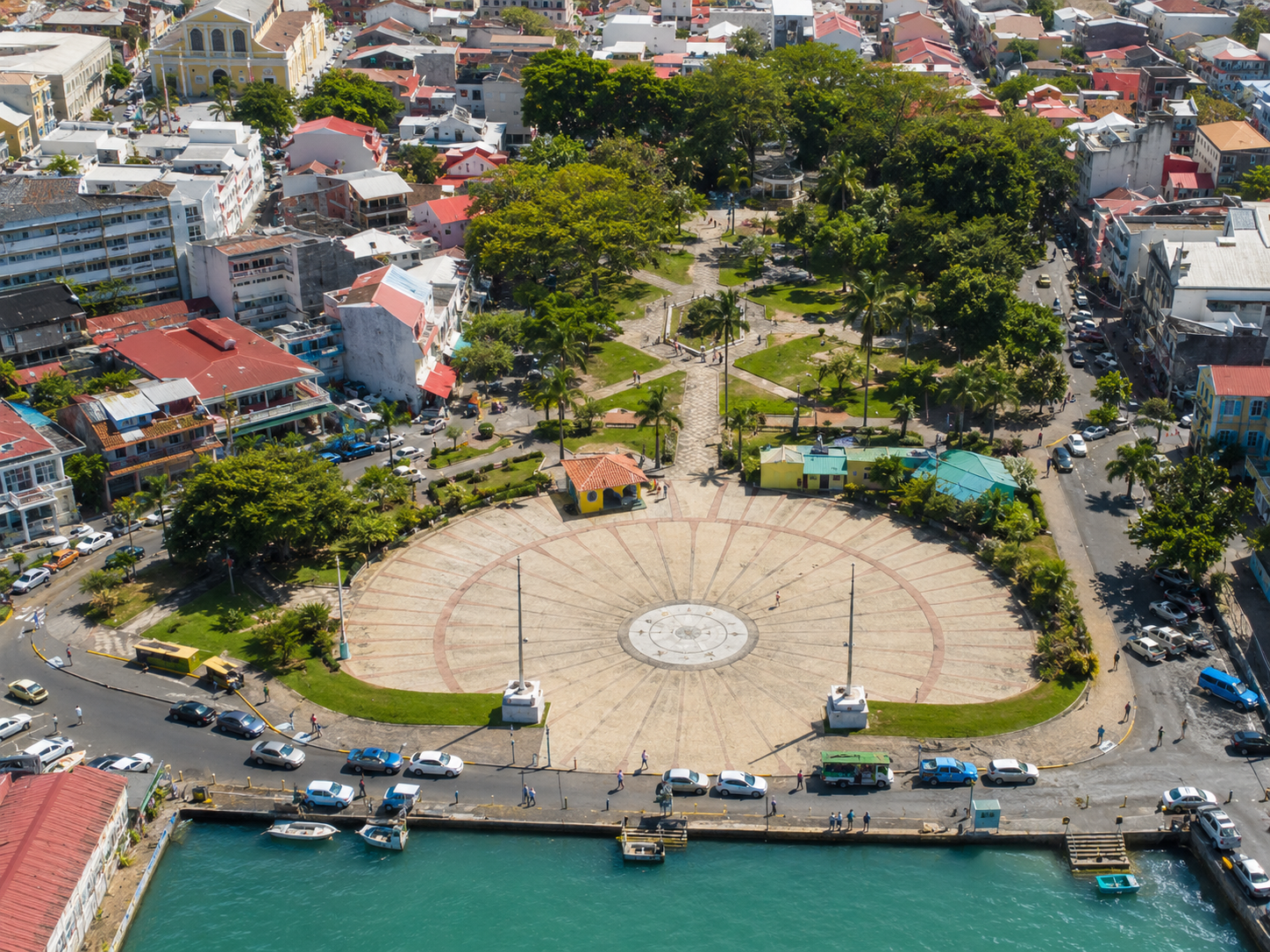 Vue aérienne plongeante de la Place de la Victoire à Pointe-à-Pitre en Guadeloupe, montrant une grande esplanade pavée en demi-cercle face à la mer, prolongée par un parc ombragé d'arbres et bordée par des rues animées et des bâtiments urbains.