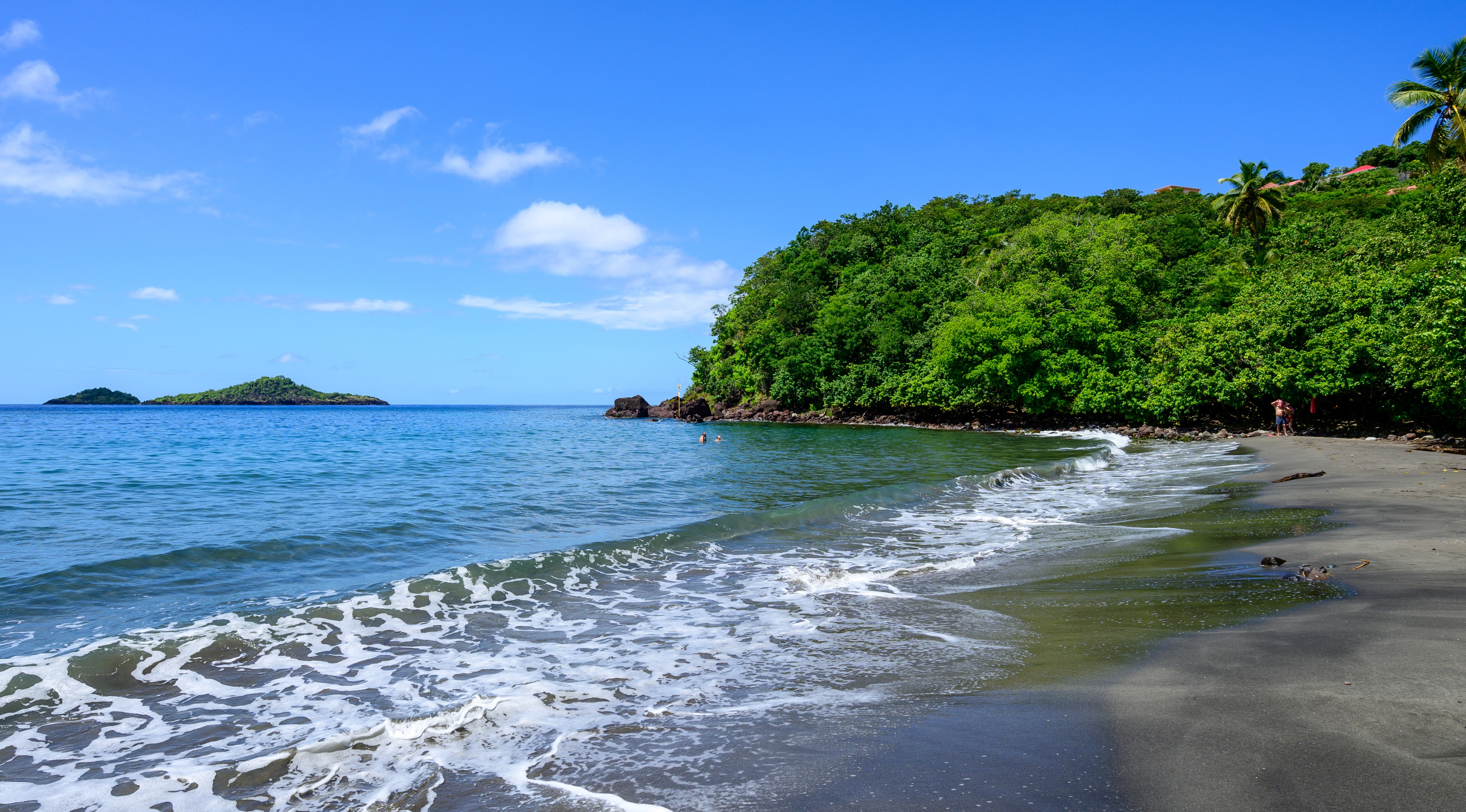 La plage de sable noir de Malendure à Bouillante en Guadeloupe, montrant de petites vagues à l'écume blanche touchant le bord, une colline verdoyante des arbres tropicaux à droite, les îlets Pigeon sur l'horizon de la mer des Caraïbes sous un ciel bleu.