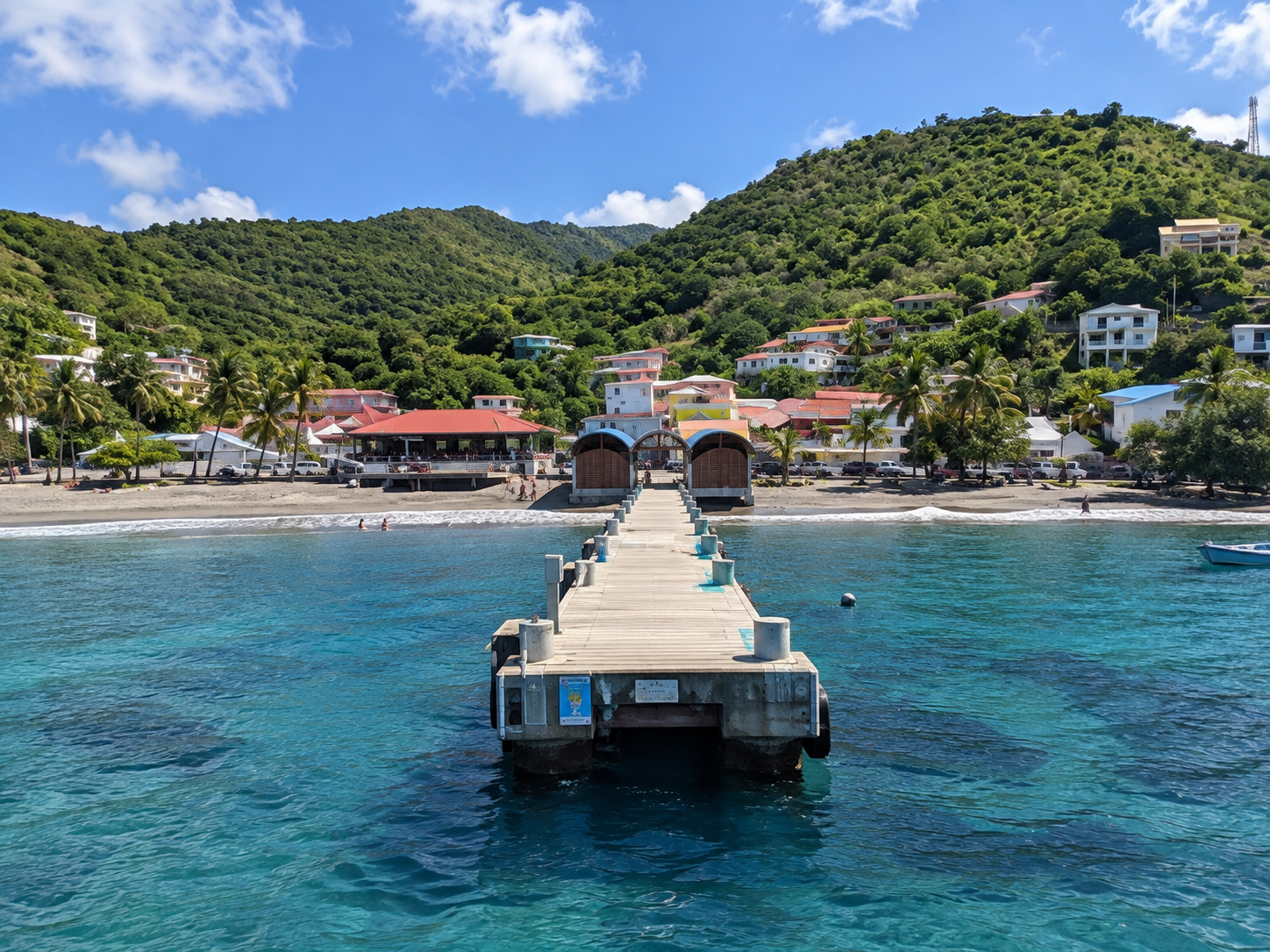 Ponton de Bellefontaine en Martinique, vue de face sur la jetée, les eaux turquoise, la plage et le village entouré de collines verdoyantes.