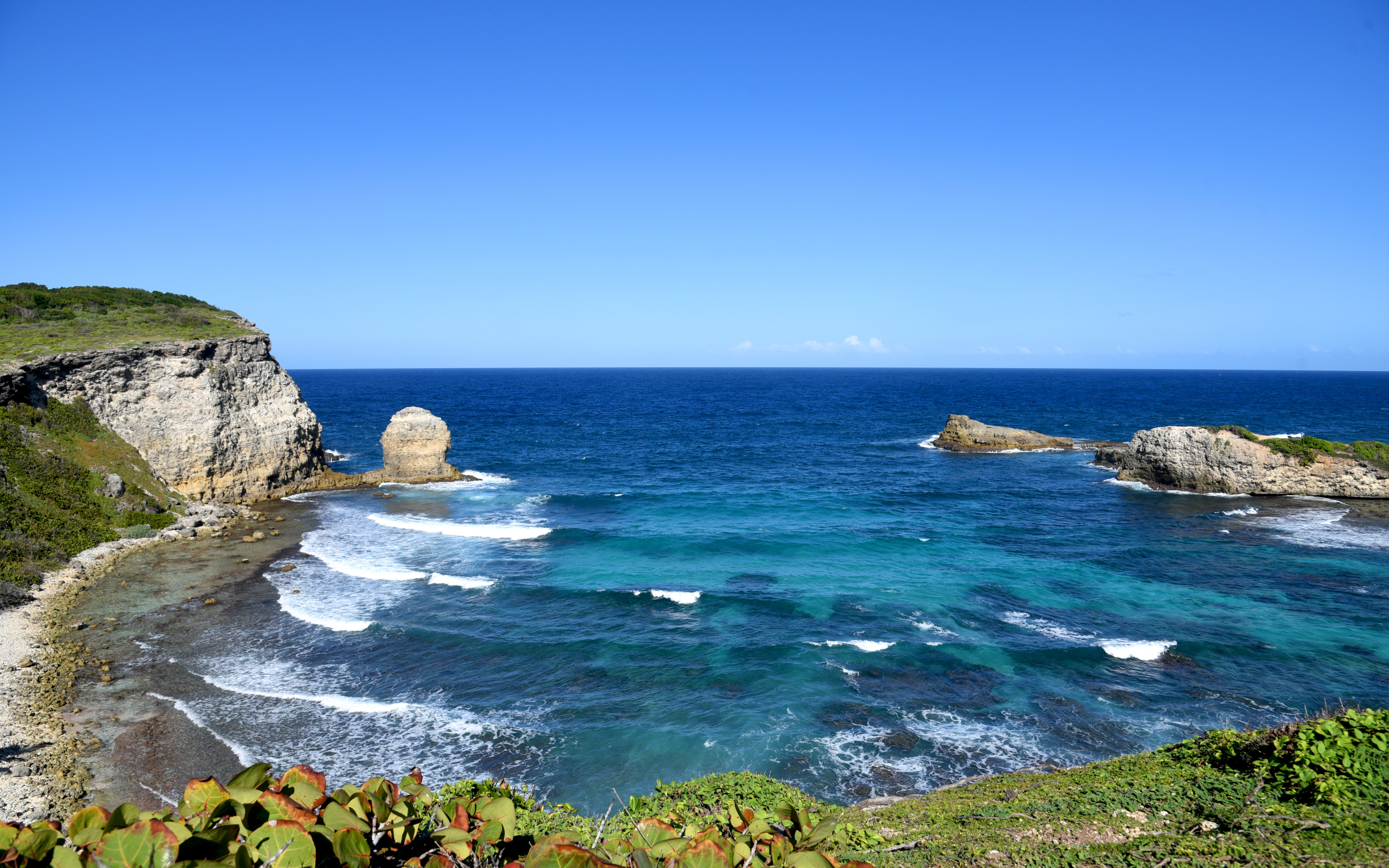 Vue panoramique de la Porte d'Enfer au Moule en Guadeloupe, montrant une baie aux eaux turquoise et agitées, entourée de falaises rocheuses sous un ciel bleu dégagé.