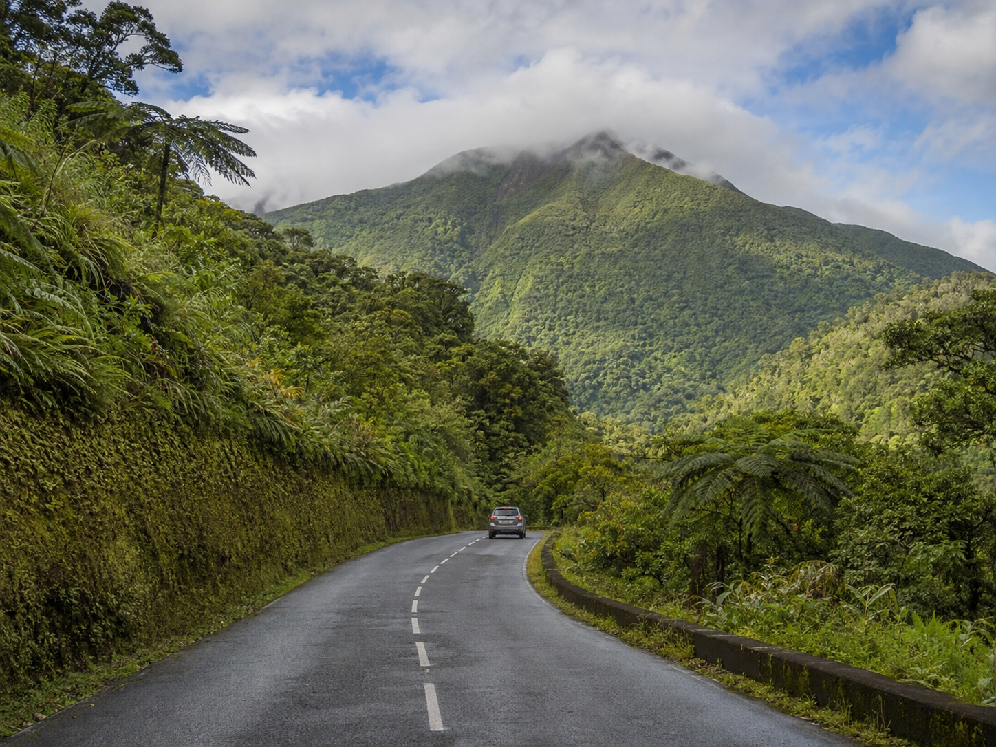 Route asphaltée sinueuse grimpant à travers une forêt tropicale dense et verdoyante, avec une voiture au loin se dirigeant vers le sommet brumeux du volcan de la Soufrière en Guadeloupe.