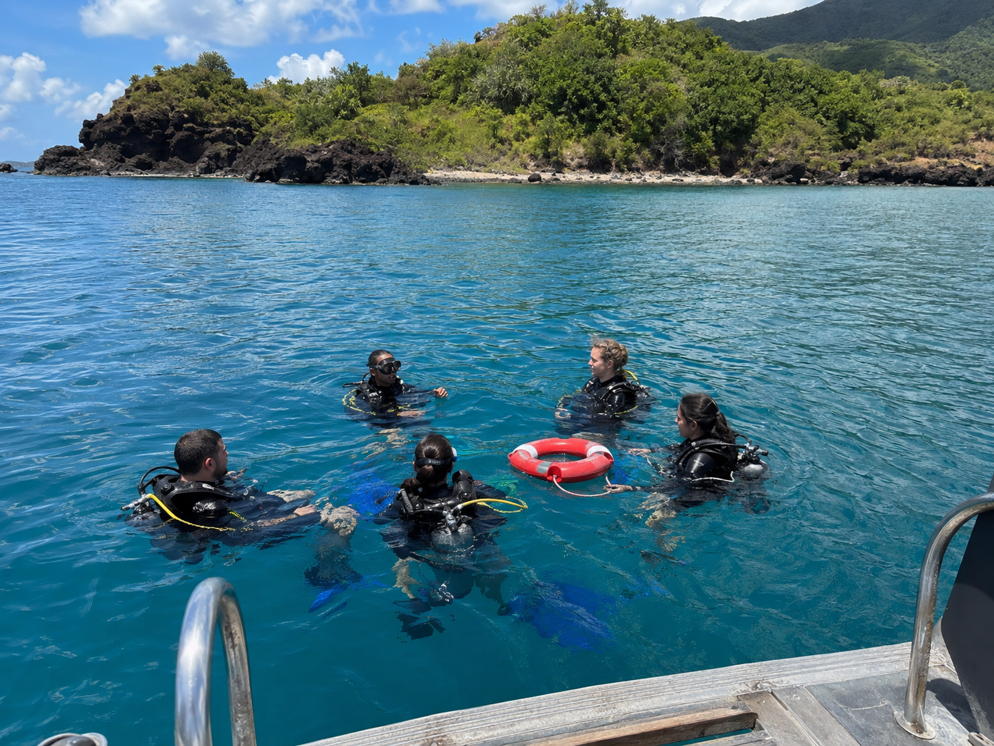 Groupe de plongeurs équipés de combinaisons et de bouteilles flottant en cercle dans l'eau turquoise de la Réserve Cousteau à Bouillante en Guadeloupe, à proximité d'une bouée de sauvetage rouge et blanche avec un îlet rocheux et boisé en arrière-plan.