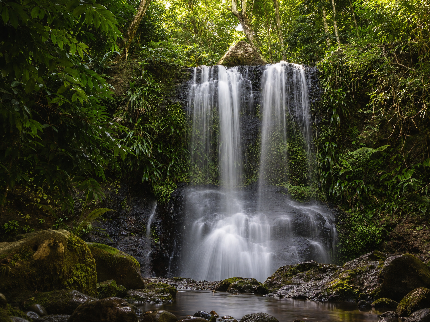 Chute d'eau du Saut des Trois Cornes à Sainte-Rose en Guadeloupe, montrant un flot vertical d'eau vive se déversant sur des roches noires dans un bassin calme, entouré d'une végétation forestière tropicale humide et de rochers moussus au premier plan.