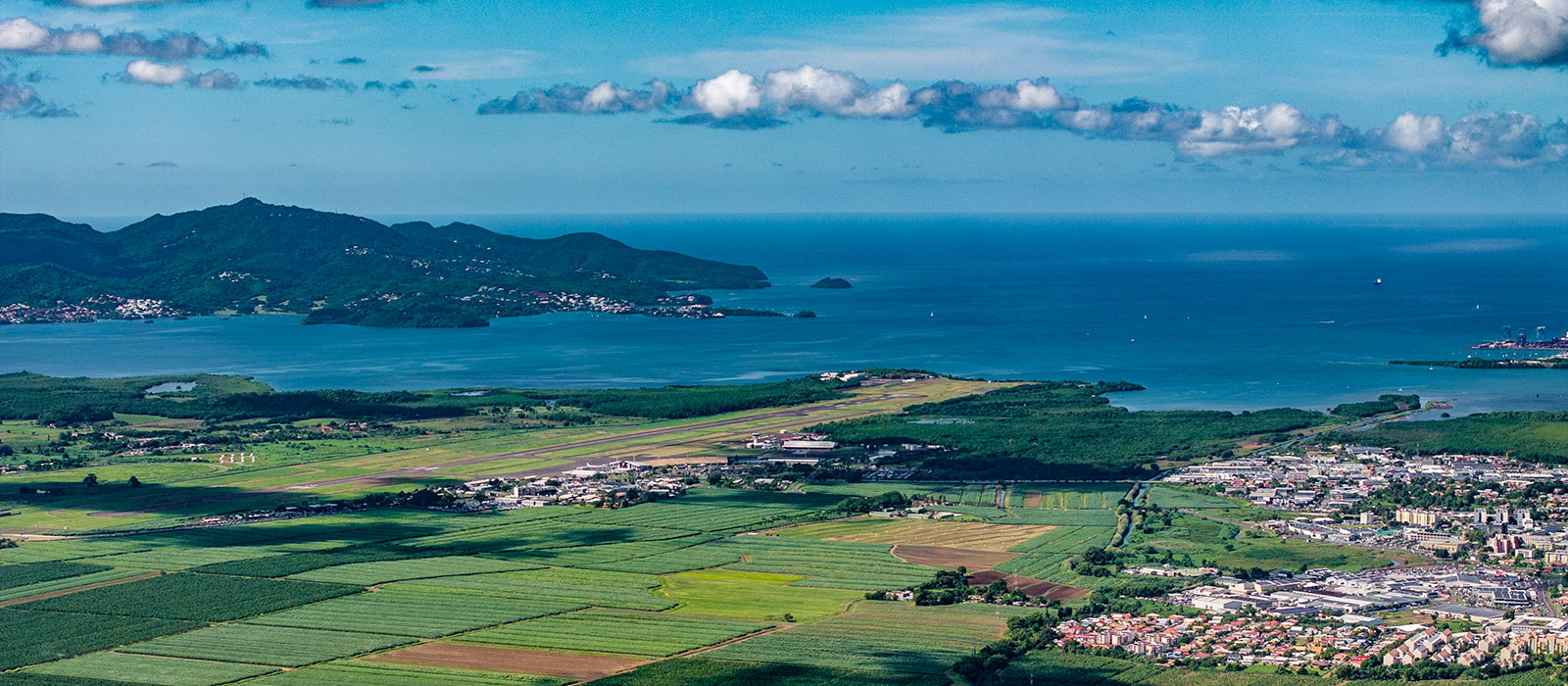 Vue aérienne de l'aéroport de Martinique Aimé Césaire : piste entre champs verts, mer des Caraïbes et collines au loin. 