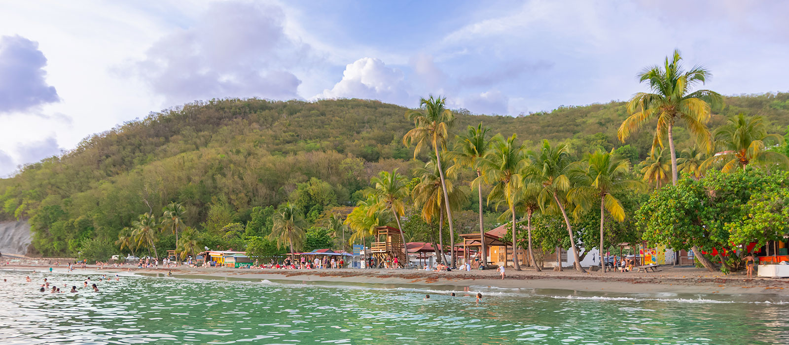 Vue depuis l'eau de la plage de Malendure à Bouillante en Guadeloupe, montrant une étendue de sable noir volcanique bordée de cocotiers, de carbets et de baigneurs, avec des collines boisées en arrière-plan sous un ciel nuageux.