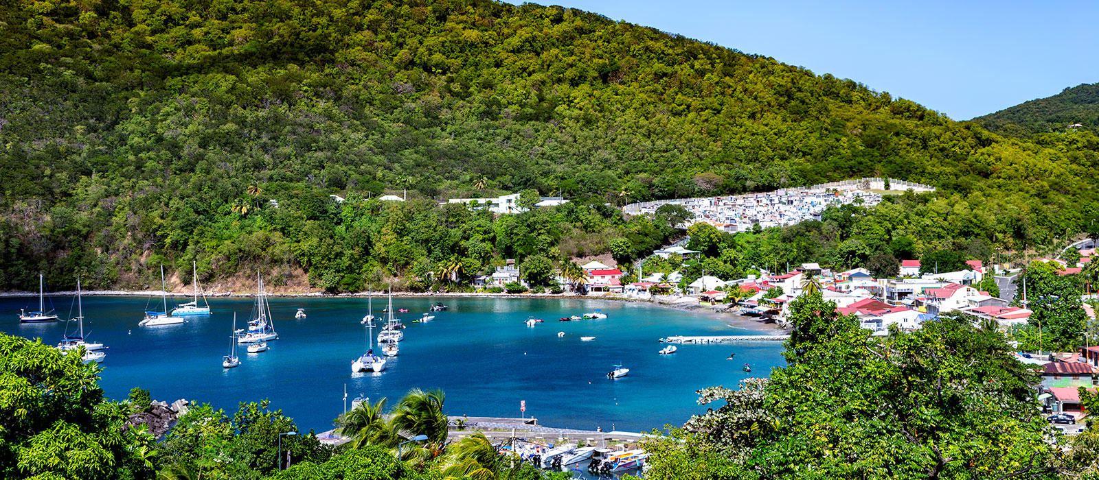 Vue en plongée de la baie de Deshaies en Guadeloupe, montrant de nombreux voiliers ancrés dans une eau bleue calme, un village côtier aux toits rouges au bord de l'eau, et le cimetière communal à damier noir et blanc flanqué sur la colline verdoyante en a