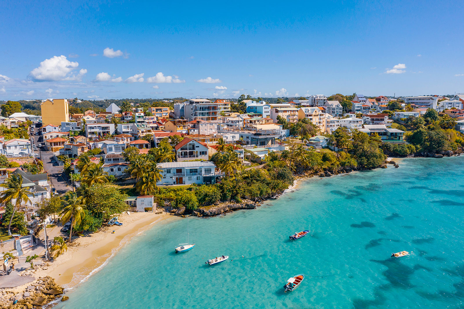 Vue aérienne du littoral de la commune du Gosier en Guadeloupe, montrant la plage de la Datcha au sable clair, plusieurs petits bateaux amarrés dans une eau turquoise transparente, et les habitations ainsi que la végétation tropicale bordant la côte roche