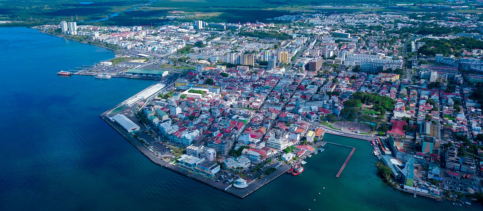 Large vue aérienne panoramique de la ville de Pointe-à-Pitre en Guadeloupe, montrant la densité urbaine des bâtiments aux toits colorés, les quais du port de croisière s'avançant dans une mer bleu foncé, et les côtes environnantes s'étendant à l'horizon.