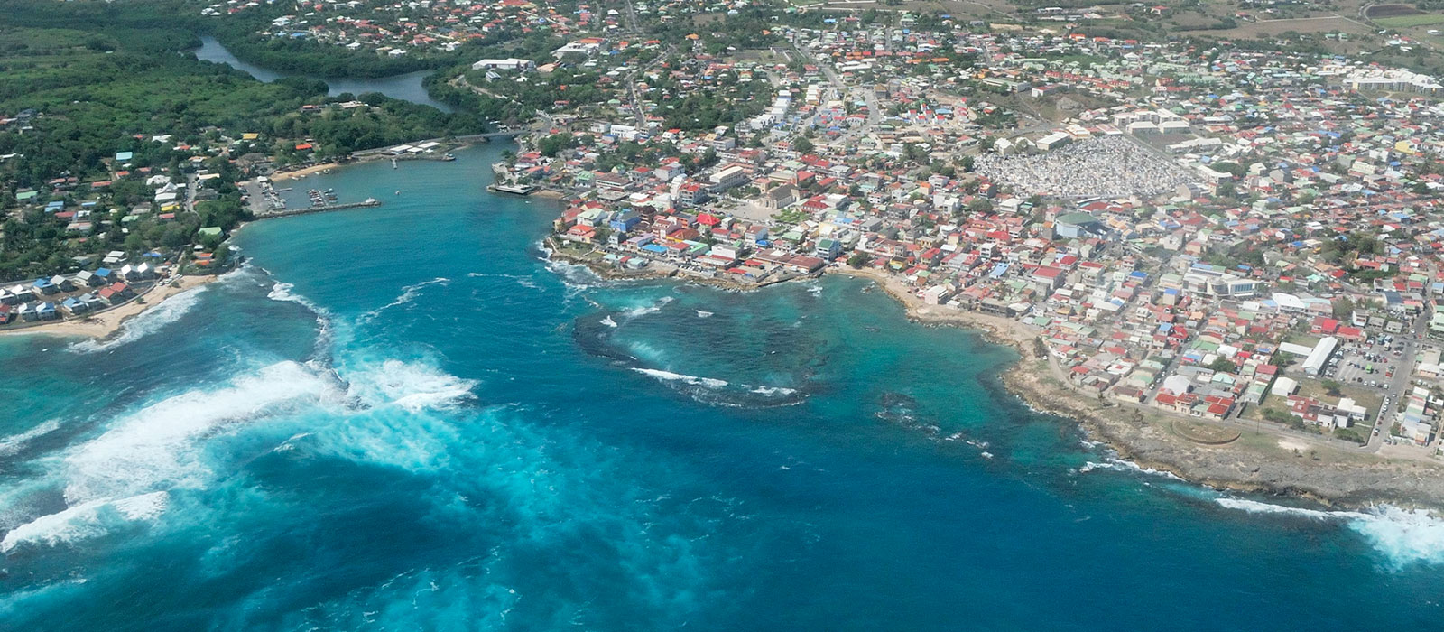 Vue aérienne du Moule en Guadeloupe : baie d'eau bleu turquoise, vagues à écume blanche et bourg littoral dense.