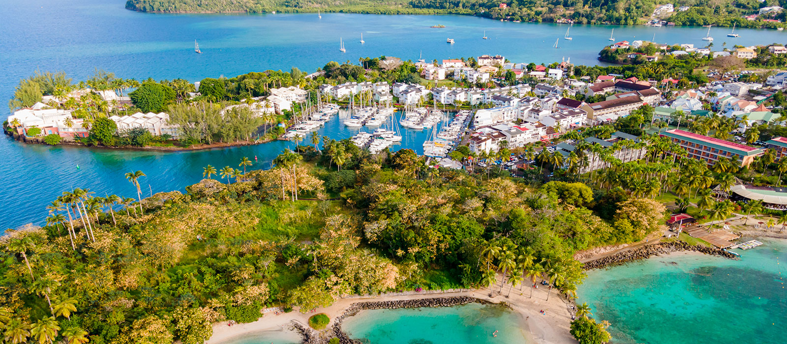 Vue aérienne de la Pointe du Bout aux Trois-Îlets : marina fortifiée, plages de sable blanc, eau turquoise et végétation. 